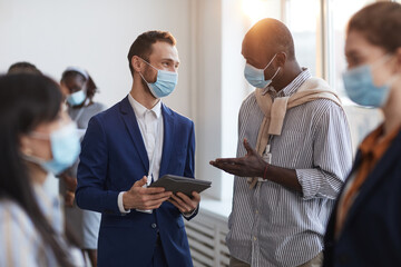 Waist up portrait of group of people chatting during break at business conference, focus on two men wearing masks with lens flare