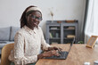 © Seventyfour - Portrait of young African-American woman using laptop at desk and looking at camera while working from home in apartment interior, copy space