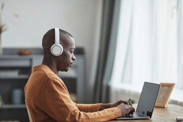 Side view portrait of African-American man using laptop at desk while working from home in minimal apartment interior, copy space