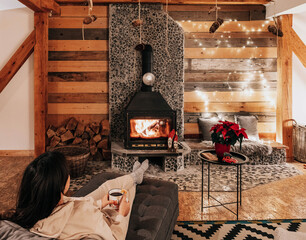 Woman is sitting with a cup of hot drink and a book near the fireplace