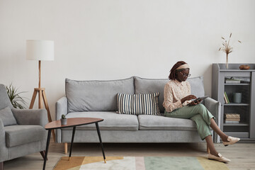 Wall Mural - Wide angle portrait of young African-American woman using digital tablet while sitting on sofa at home in minimal grey interior, copy space