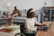 © Seventyfour - Portrait of young African-American woman using wheelchair while working from home in minimal grey interior, copy space