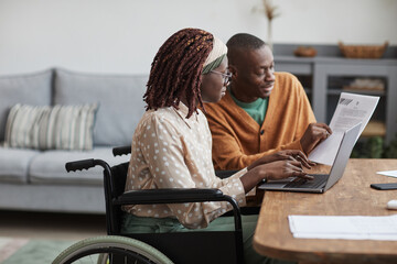 Side view portrait of young African-American woman using wheelchair working from home with husband helping her copy space