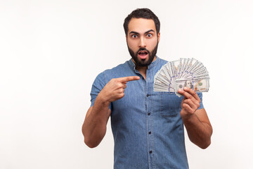 Surprised bearded man pointing at fan of dollar cash in his hand, money exchange, shocked with interest rate. Indoor studio shot isolated on white background