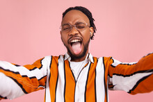 Extremely Happy Bearded Man Blogger With Dreadlocks Showing Tongue Out, Having Fun Posing On Selfie Camera, Live Streaming. Indoor Studio Shot Isolated On Pink Background