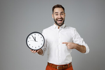 Excited funny young bearded business man 20s wearing classic white shirt standing pointing index finger on clock isolated on grey color background studio portrait. Achievement career wealth concept.