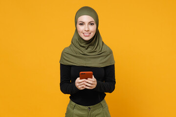 Wall Mural - Smiling young arabian muslim woman wearing hijab black green clothes using mobile cell phone typing sms message isolated on yellow color background studio portrait. People religious lifestyle concept.