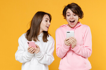 Wall Mural - Excited amazed smiling two young women friends 20s wearing casual white pink hoodies standing using mobile cell phone typing sms message isolated on bright yellow color background studio portrait.