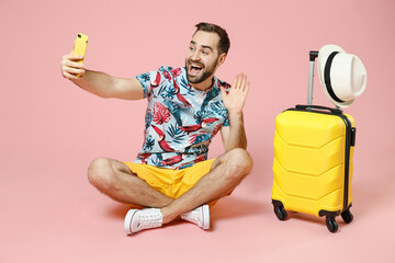 Wall Mural - Full length excited young traveler tourist man sit on floor doing selfie shot on mobile phone greeting with hand isolated on pink background. Passenger travel on weekend. Air flight journey concept.