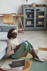 Wall Mural - Vertical side view portrait of modern African-American woman working from home while sitting on floor at graphic carpet and reading documents, copy space