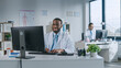 © Gorodenkoff - Happy Smiling African American Medical Doctor is Working on a Computer in a Health Clinic. Physician in White Lab Coat is Browsing Medical History Behind a Desk in Hospital Office.