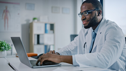 Happy African American Family Medical Doctor is Working on a Laptop Computer in a Health Clinic. Physician in White Lab Coat is Browsing Medical History Behind a Desk in Hospital Office. 