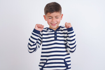 little cute boy kid wearing stripped t-shirt against white wall rejoicing his success and victory clenching fists with joy being happy to achieve aim and goals. Positive emotions, feelings.