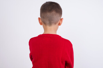 Little cute boy kid wearing red knitted sweater against white wall standing backwards looking away with arms on body.