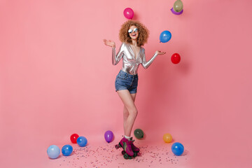 Wall Mural - Adorable girl in sunglasses, silver blouse and shorts roller skating in pink studio among multicolored balloons