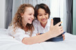 © DragonImages - Cheerful multi-ethnic young lesbian couple lying on bed and taking selfie on smartphone
