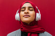 © David Munoz/ADDICTIVE STOCK - Dreamy Arab female in wireless headphones and traditional hijab standing near urban red building and enjoying music with closed eyes