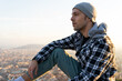 © David Munoz/ADDICTIVE STOCK - Side view of thoughtful hipster male traveler in warm checkered hoodie and hat looking away while sitting on hill against blurred town in autumn day