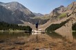 © German Linares/ADDICTIVE STOCK - Back view of male explorer standing on tree trunk in clear pond and enjoying view of majestic mountainous landscape in summer