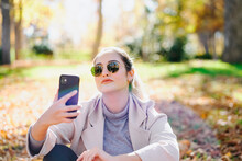 Female In Sunglasses Sitting In Autumn Park And Texting On Smartphone While Enjoying Conversation And Looking Away