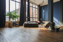 Dark Interior Of A Modern Stylish Huge Open-plan Loft-style Studio Apartment With Columns And High Ceilings. Dark Blue Primed Walls Are Decorated With Wood. Sunlight Enters Through Huge Windows.