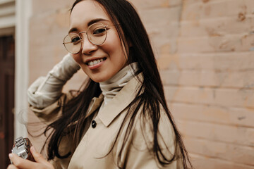 Wall Mural - Asian woman looking at camera. Girl in beige trench coat posing against brick wall
