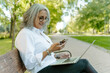 © Jake Jakab/ADDICTIVE STOCK - Side view of mature female entrepreneur sitting on bench in park and browsing smartphone while reading working messages