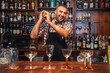 © Javier De La Torre/ADDICTIVE STOCK - Excited middle aged mixologist in checkered shirt and apron looking at camera and mixing alcohol drink in shaker while standing behind counter with glasses in bar
