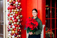 Woman Standing Outside Shop Red Door With Christmas Decorative Baubles Holding Pot Of Poinsettia Flower In Daylight Looking At Camera