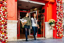 Woman Walking Outside Shop With Female Friend At Red Door With Christmas Decorative Baubles Holding Flower Bouquet