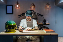 Front View Of A Man Writing In A Notebook On A Wooden Table Next To A Motorcycle Helmet