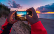 © Javier Martinez/ADDICTIVE STOCK - Back view of unrecognizable female traveler with long dark hair in casual clothes taking picture of amazing sunset over sea with sandy beach on smartphone during holidays in Cantabria