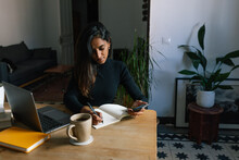 Indian Female Entrepreneur Sitting At Table And Writing Plans In Notebook While Working At Home In Cozy Workplace