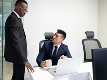 Serious Young Asian Male Boss In Formal Suit With Cup Of Coffee Pointing At Laptop Screen And Asking African American Manager To Explain Problem Solution During Meeting In Office