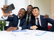 © Jose Carlos Cerdeno/ADDICTIVE STOCK - Group of smiling multiracial male business colleagues in formal suits gathering near table with diagrams and taking selfie on mobile phone during meeting in modern office