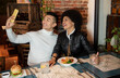 © Jose Carlos Cerdeno/ADDICTIVE STOCK - Happy young Asian man and African American girlfriend having fun and taking selfie on smartphone during lunch in restaurant