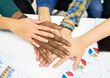 © Jose Carlos Cerdeno/ADDICTIVE STOCK - From above of crop anonymous multiracial colleagues in casual clothes gathering around table with charts and stacking hands together while working on business development plan in workplace