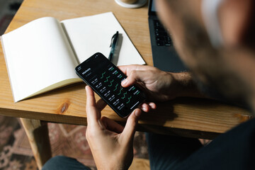 High angle of crop anonymous male entrepreneur using mobile app and working with diagrams at home