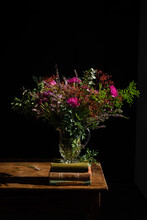 Bunch Of Assorted Aromatic Wildflowers Placed In Glass Vase On Rustic Wooden Table With Pile Of Old Books On Black Background In Studio