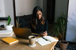 © Joy Zamora/ADDICTIVE STOCK - Female sitting at table with cup of coffee and browsing smartphone while working on online project