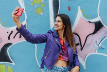 Smiling Trendy Millennial Woman In Stylish Wear Standing Near Colorful Painted Graffiti Wall And Taking Selfie On Smartphone