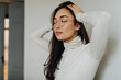 © Look! - Brunette girl in glasses and sweater poses against white wall in apartment and massages her head