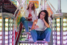Young Female Friends In Casual Clothes With Candies In Hands Looking At Camera While Sitting On Steps Of Attraction In Funfair In Summer Evening