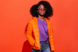© Manuel Ruiz/ADDICTIVE STOCK - Young black female in purple shirt with denim and orange jacket standing with hands in pockets on red background looking at camera