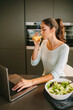 © Luis Tenza/ADDICTIVE STOCK - Side view of young female freelancer working on project on laptop while drinking orange juice and having lunch in kitchen at home