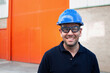 © Manuel Ruiz/ADDICTIVE STOCK - Positive confident adult male workman in blue protective hardhat and eyeglasses smiling friendly while standing against bright orange wall of workshop