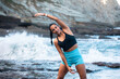 © Pablo Garcia/ADDICTIVE STOCK - Positive female athlete warming up body and doing side bends while stretching arms before workout on seashore and looking at camera