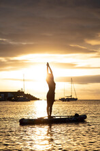 Side View Silhouette Of Slim Female Standing In Mountain Pose With Arms Up While Practicing Yoga On Floating Paddle Board Against Sunset Sky