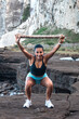 © Pablo Garcia/ADDICTIVE STOCK - Delighted athletic female in sportswear doing squat exercise with wooden stick above head while training at seaside and looking at camera