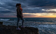 © Oscar De Dios/ADDICTIVE STOCK - Side view of African American female in warm clothes standing on rocky shore near waving sea and admiring amazing sundown